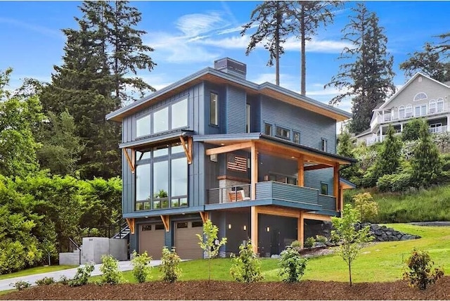 rear view of property featuring a balcony, driveway, an attached garage, a chimney, and a lawn