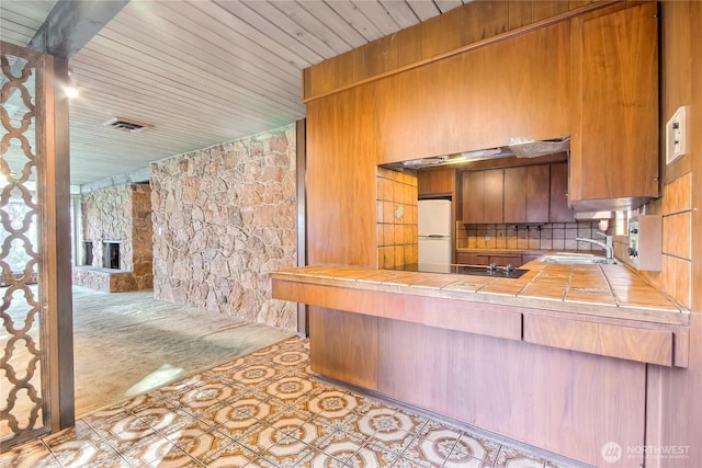 kitchen with tile counters, brown cabinets, freestanding refrigerator, black electric stovetop, and a sink