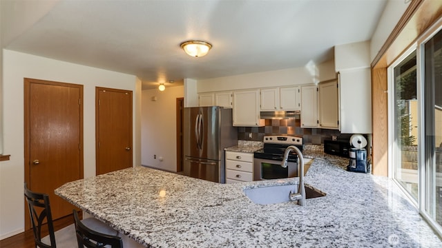 kitchen with light stone counters, under cabinet range hood, a peninsula, a sink, and appliances with stainless steel finishes