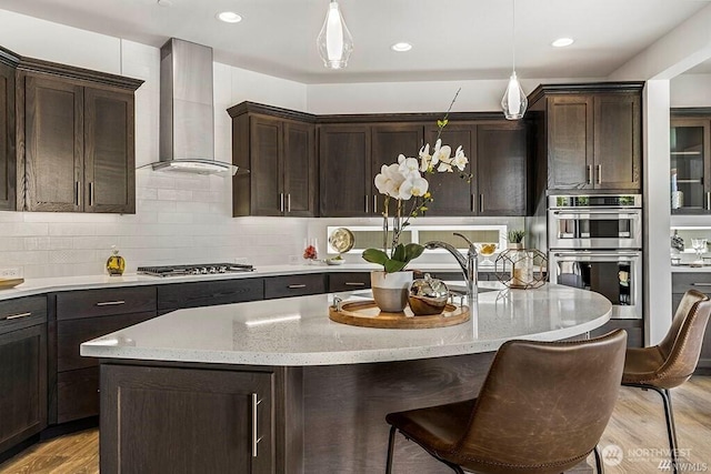 kitchen with appliances with stainless steel finishes, hanging light fixtures, wall chimney exhaust hood, light stone countertops, and a kitchen island