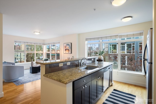 kitchen with a center island with sink, light stone countertops, stainless steel appliances, dark cabinetry, and a sink