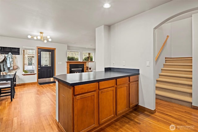 kitchen with brown cabinetry, a lit fireplace, light wood-style floors, dark countertops, and a notable chandelier