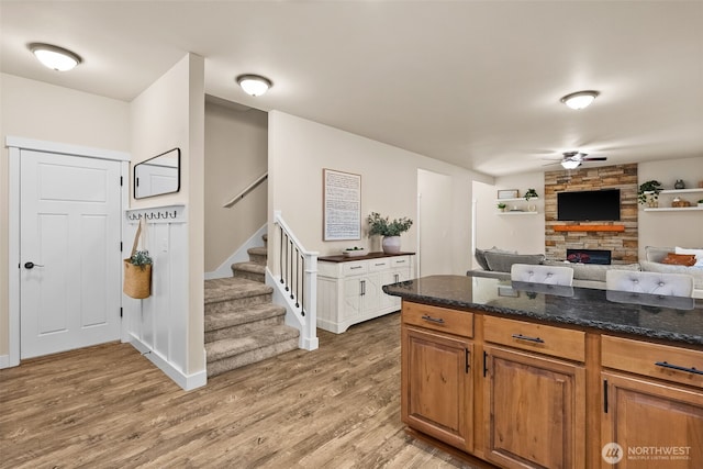 kitchen with brown cabinets, a fireplace, light wood finished floors, a ceiling fan, and dark stone countertops