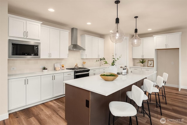 kitchen featuring a center island with sink, stainless steel appliances, white cabinets, wall chimney range hood, and wood finished floors