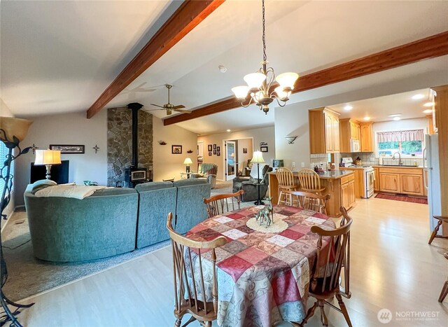 dining space with lofted ceiling with beams, ceiling fan with notable chandelier, a wood stove, and light wood-style floors