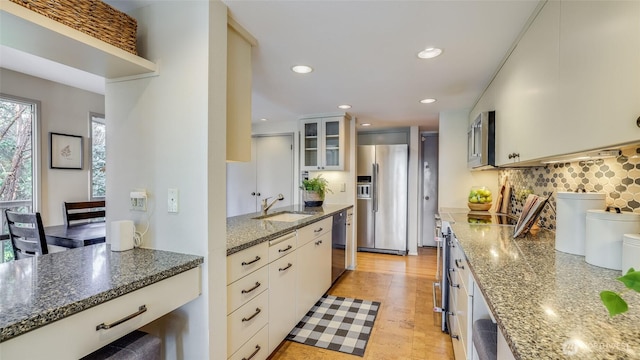 kitchen with tasteful backsplash, glass insert cabinets, appliances with stainless steel finishes, dark stone countertops, and a sink