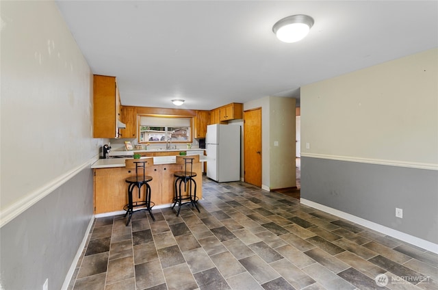 kitchen with light countertops, a peninsula, freestanding refrigerator, brown cabinetry, and a sink