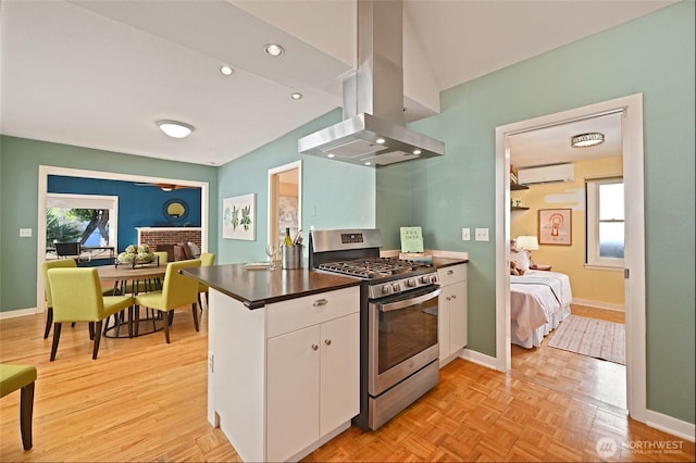 kitchen with island exhaust hood, dark countertops, a wall mounted AC, stainless steel gas stove, and white cabinetry