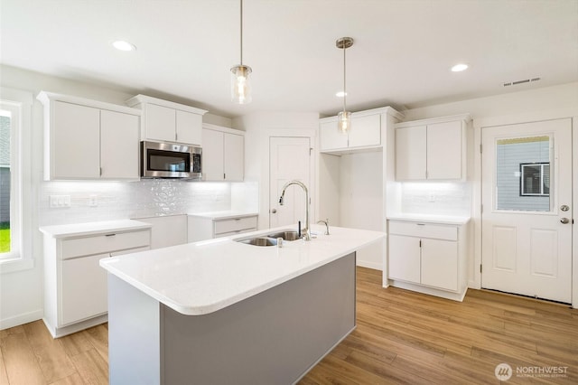 kitchen with a sink, an island with sink, light wood-style flooring, stainless steel microwave, and backsplash