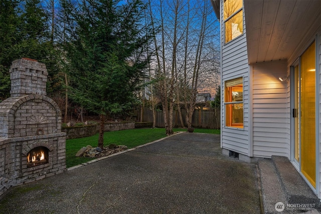 patio terrace at dusk featuring a warm lit fireplace, a lawn, and fence