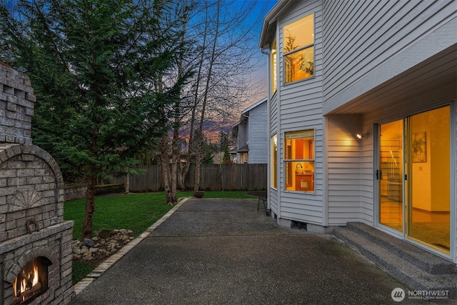 patio terrace at dusk with a fenced backyard, a yard, and a lit fireplace