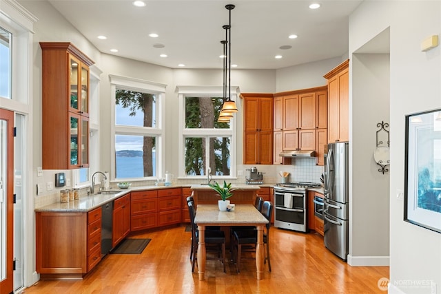 kitchen featuring under cabinet range hood, hanging light fixtures, appliances with stainless steel finishes, light stone countertops, and glass insert cabinets