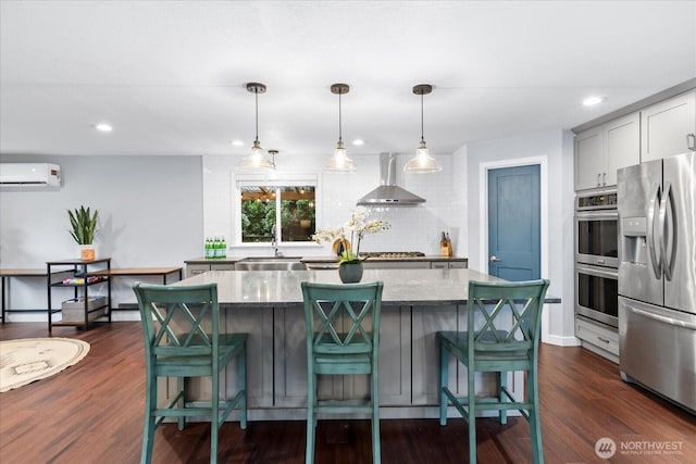 kitchen with pendant lighting, appliances with stainless steel finishes, an AC wall unit, light stone countertops, and wall chimney exhaust hood