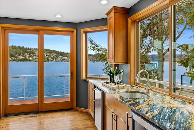 interior space featuring light wood finished floors, brown cabinetry, dark stone countertops, a water view, and a sink