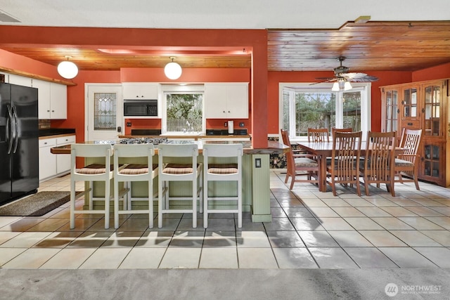 kitchen with wooden ceiling, a breakfast bar area, black refrigerator with ice dispenser, white cabinetry, and light tile patterned flooring