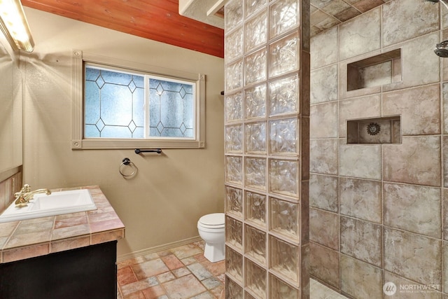 bathroom with baseboards, toilet, walk in shower, stone tile flooring, and vanity