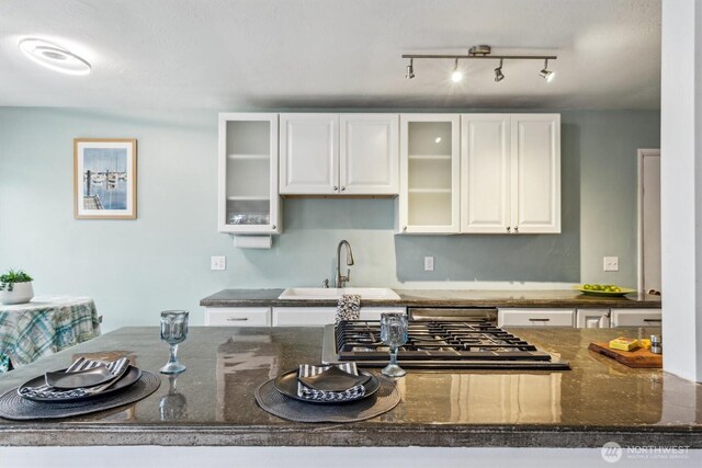 kitchen with stainless steel gas cooktop, glass insert cabinets, dark stone countertops, white cabinetry, and a sink