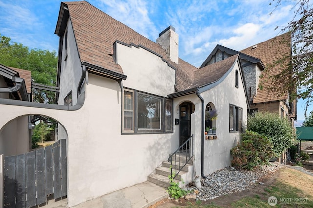 tudor-style house featuring entry steps, fence, a chimney, and stucco siding