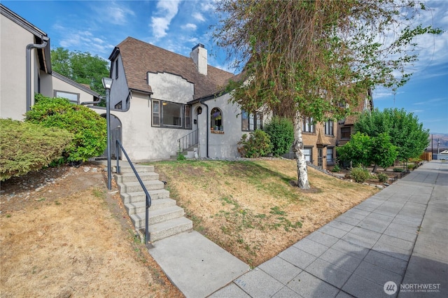 tudor house with entry steps, stucco siding, a chimney, roof with shingles, and a front yard