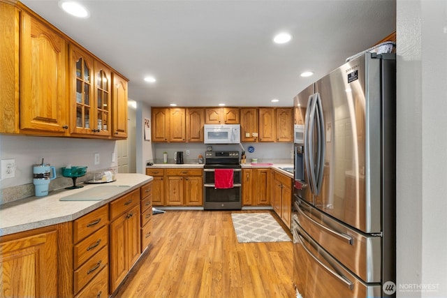 kitchen with light countertops, appliances with stainless steel finishes, light wood-type flooring, brown cabinets, and glass insert cabinets