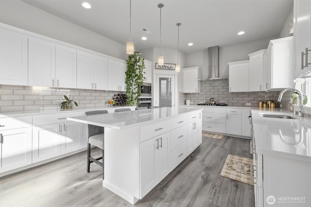 kitchen featuring a kitchen island, a sink, white cabinetry, light countertops, and wall chimney exhaust hood