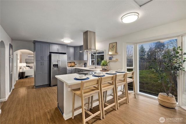 kitchen with arched walkways, gray cabinets, island range hood, a peninsula, and stainless steel fridge with ice dispenser