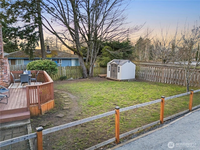 yard at dusk featuring a deck, outdoor dining area, a fenced backyard, an outdoor structure, and a storage unit