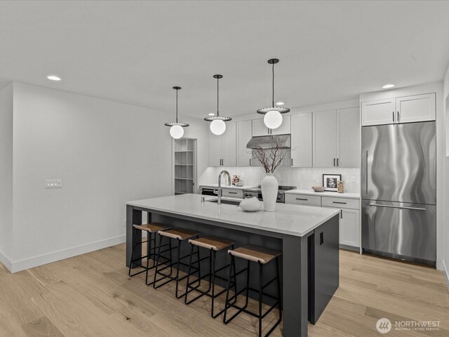 kitchen featuring a kitchen island with sink, a sink, white cabinets, light countertops, and stainless steel fridge