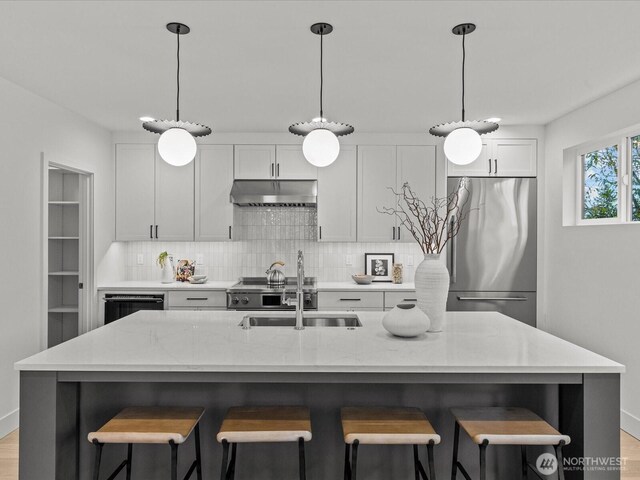 kitchen featuring light stone counters, stainless steel refrigerator, a center island with sink, and under cabinet range hood