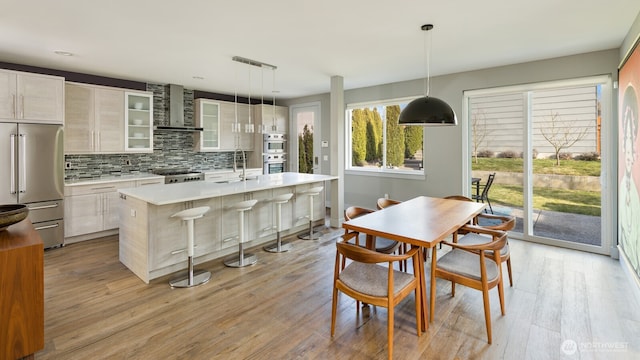 kitchen featuring stainless steel appliances, light countertops, glass insert cabinets, a kitchen island with sink, and wall chimney exhaust hood