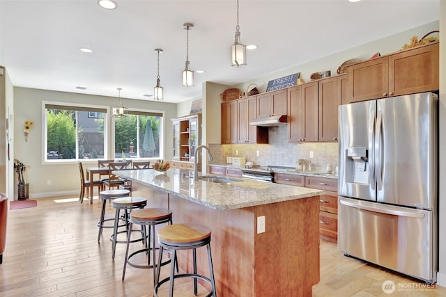 kitchen featuring light stone counters, stainless steel appliances, a sink, brown cabinetry, and a center island with sink