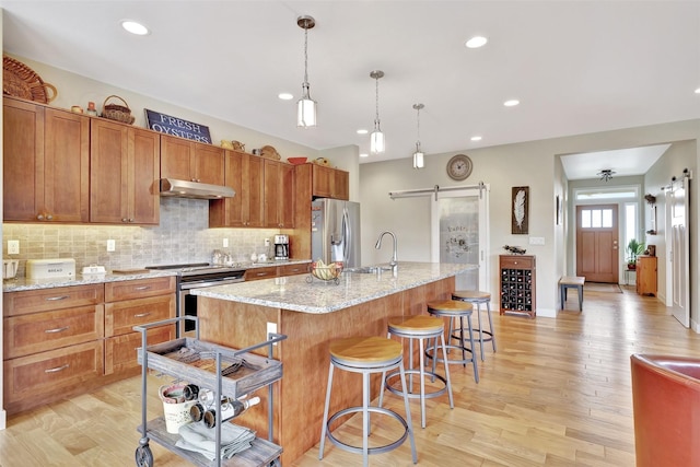 kitchen with a barn door, under cabinet range hood, appliances with stainless steel finishes, brown cabinets, and a center island with sink