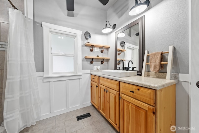 bathroom with tile patterned flooring, a decorative wall, a ceiling fan, vanity, and wainscoting