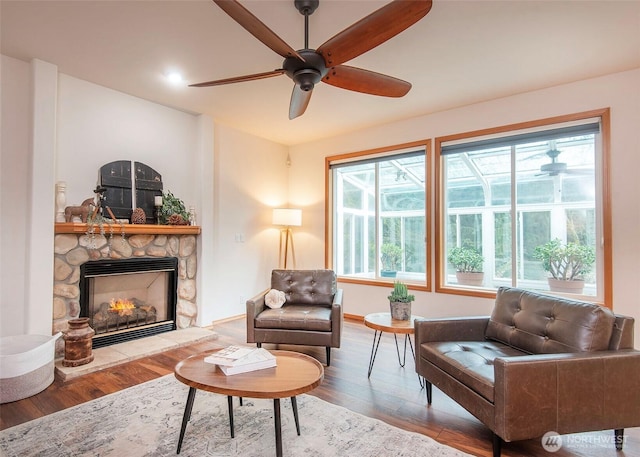 living area with a stone fireplace, wood finished floors, a ceiling fan, and baseboards