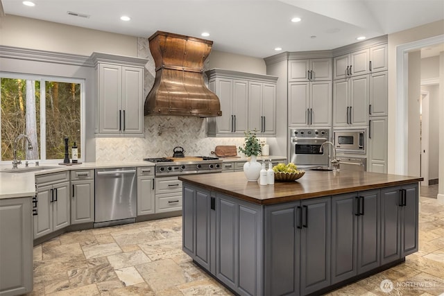 kitchen with gray cabinetry, butcher block counters, a sink, appliances with stainless steel finishes, and custom range hood