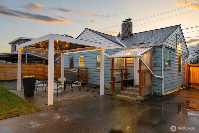 back of property at dusk featuring a shingled roof, a chimney, a patio area, and fence