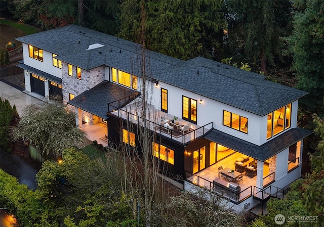 rear view of house featuring a balcony, stone siding, an attached garage, and roof with shingles