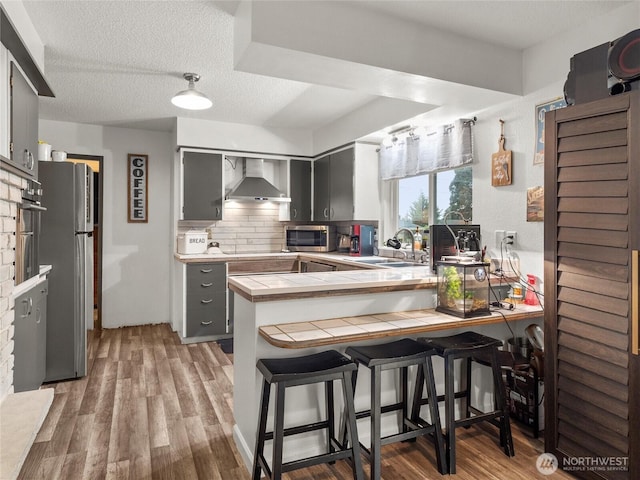 kitchen with gray cabinetry, a peninsula, a sink, tile counters, and wall chimney exhaust hood