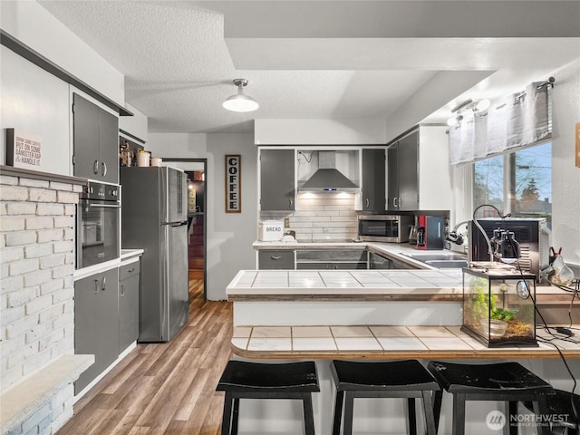 kitchen featuring tile countertops, stainless steel appliances, a breakfast bar, a sink, and wall chimney exhaust hood
