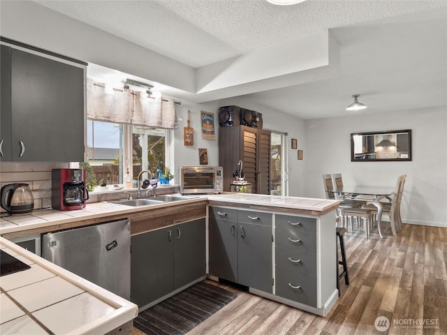 kitchen with tile countertops, a sink, wood finished floors, a peninsula, and dishwashing machine