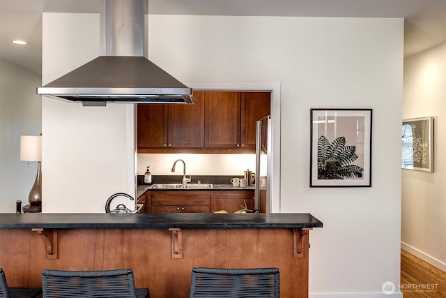 kitchen featuring a breakfast bar area, dark countertops, a sink, island range hood, and a peninsula