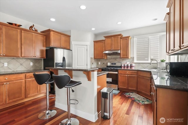 kitchen with under cabinet range hood, a sink, a kitchen breakfast bar, stainless steel appliances, and brown cabinetry