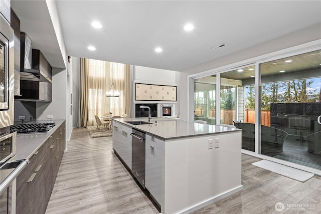 kitchen with light wood-style flooring, stainless steel appliances, a sink, modern cabinets, and a center island with sink