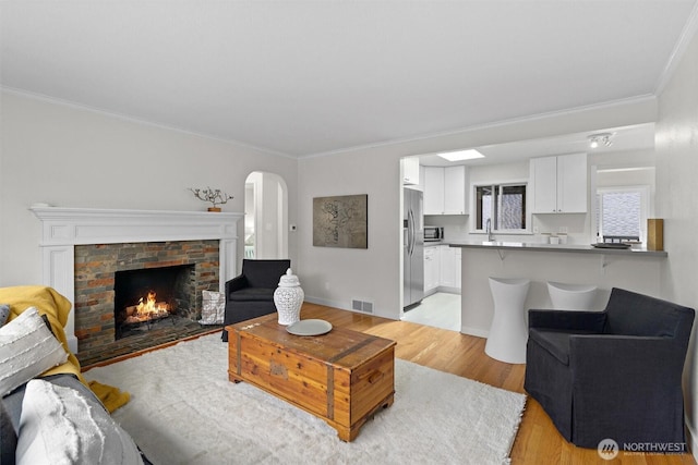 living room featuring a warm lit fireplace, visible vents, arched walkways, crown molding, and light wood-style floors