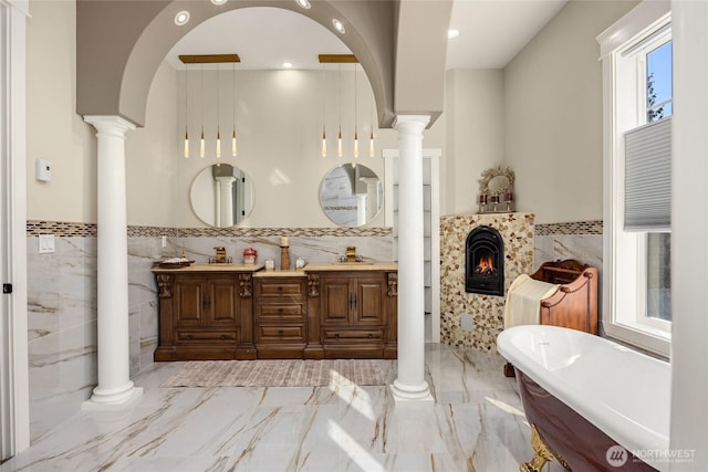 full bathroom featuring marble finish floor, a fireplace, decorative columns, a freestanding bath, and a sink