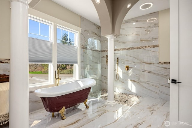 full bathroom featuring stone wall, marble finish floor, a freestanding bath, and decorative columns