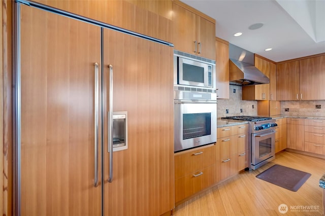 kitchen with light stone counters, backsplash, wall chimney range hood, built in appliances, and light wood-type flooring