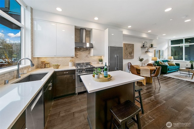 kitchen featuring a sink, high quality appliances, white cabinetry, light countertops, and wall chimney range hood