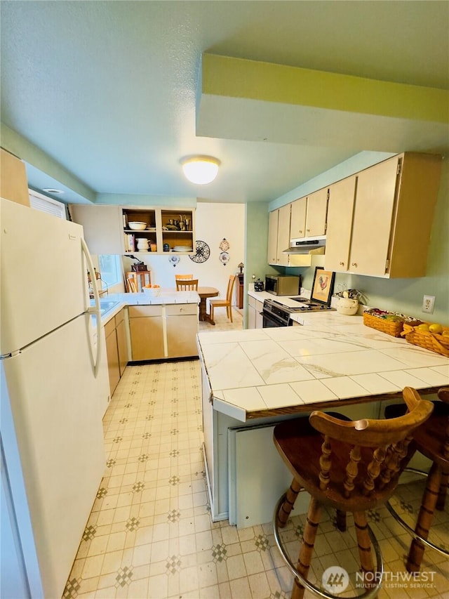 kitchen featuring tile countertops, under cabinet range hood, a peninsula, range, and freestanding refrigerator