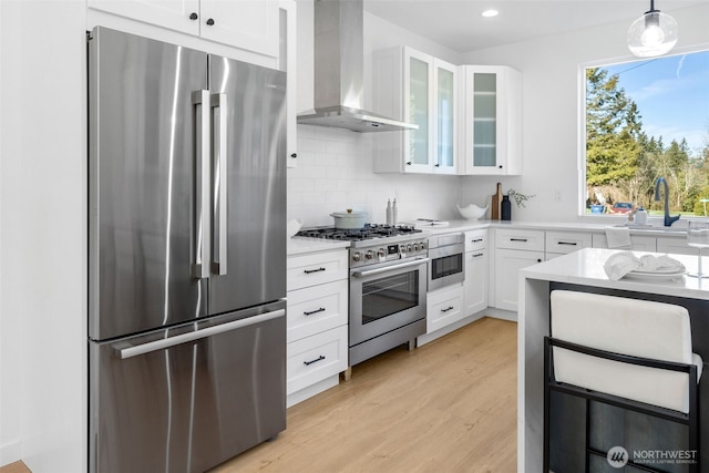 kitchen with stainless steel appliances, a sink, white cabinets, light countertops, and wall chimney range hood